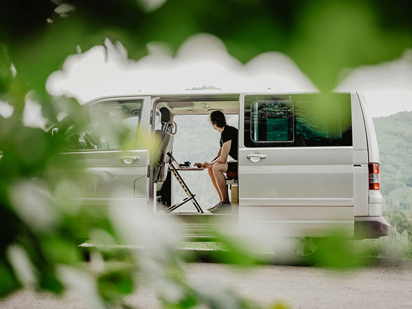 Mann sitzt in einem VW Bus und schaut in die Ferne, vor ihm steht ein Laptop auf einem Standsome